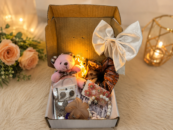Decorative box with a teddy bear, jewelry, and other items on a soft surface with flowers and candles in the background.